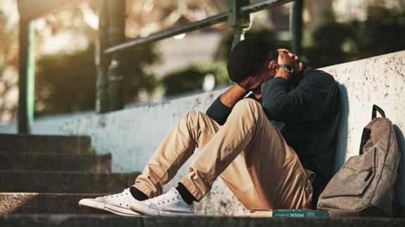 Young adult male sitting outside on the ground. His knees are bent and his hands are clasped behind his neck with his elbows on his knees. He looks upset